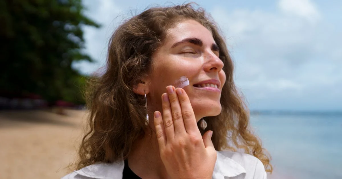 A smiling woman at the beach applying sunscreen to her face under a sunny sky.
